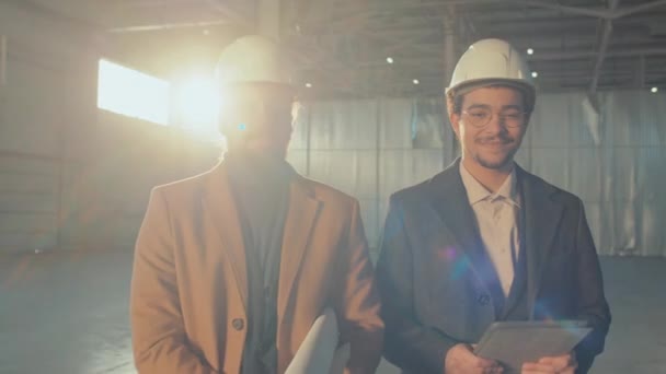 身着硬帽 头戴平板和纸卷 面带微笑的白种人和比尔卡利亚男性商业伙伴在空旷的建筑工地摆出一副中影 看着相机 — 图库视频影像