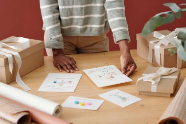 Anneler günü için el yapımı kartpostal seçen bir kızın eli beyaz kurdelelerle paketlenmiş hediye kutularıyla masanın yanında dururken...