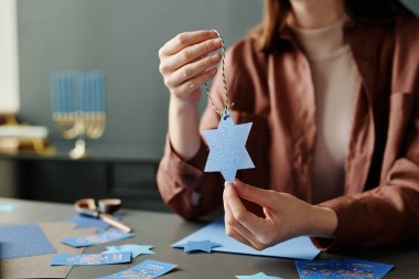Elinde mavi kağıttan kesilmiş el yapımı oyuncak hediyeler tutan genç bir kadın. Masasında Hanuka kartpostallarıyla oturuyor.