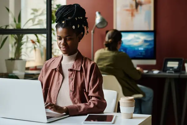 Young African American businesswoman looking at laptop screen while sitting by workplace and organizing work or carrying out task
