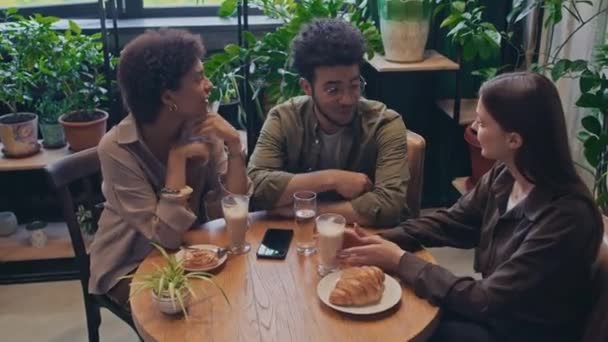 中高角镜头拍摄的年轻的白人和非洲裔美国妇女和中东男子坐在舒适的餐馆餐桌前 喝咖啡和小吃 并进行着热烈的对话 — 图库视频影像