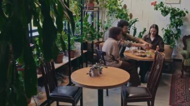 Geniş bir grup genç çoklu etnik öğrenci rahat bir restoranda yeşil bitkilerin arasında öğle yemeği yiyor, kahve içiyor, sohbet ediyor, bir şeyler tartışıyor ve gülüyor.