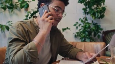 Orta boy, kıvırcık saçlı, kafede ya da birlikte çalışan, sunum kağıdındaki verilere bakan ve akıllı telefondan konferans görüşmesi yapan Orta Doğulu bir adam.