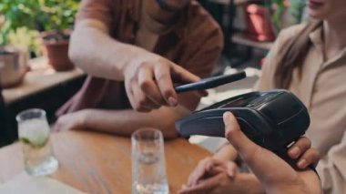 Ödeme terminalini elinde tutan tanınmayan garsonun yakın plan fotoğrafları, akıllı telefon uygulaması ile ödeme yapan kimliği belirsiz genç adam, restoranda kız arkadaşıyla masada otururken.