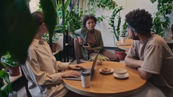坐在办公室餐厅餐桌边或同事旁边 与邻桌的非洲裔美国女同事聊天的白种人女性 带着笔记本电脑和白种人的中景照片 — 图库视频影像