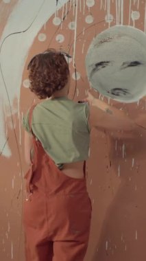 Tulum giymiş tanınmayan kadın ressamın orta dikey arka çekimi, kıvırcık saçlı, sanat stüdyosundaki duvarda ekstra büyük, soyut bir duvar resmi üzerinde çalışıyor, gri dairesel şekildeki siyah boyayı kirletiyor.