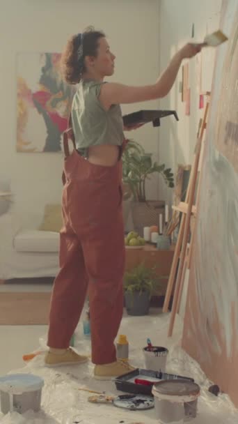 スタジオに立っているジャンプスーツの白人女性の装飾者の完全な縦のショット ペンキが付いている皿を握り ブラシが付いている合板シートかキャンバスで抽象的なラインを描きます — ストック動画