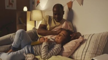 Orta boy, Afro-Amerikan bir babanın, modern, rahat bir yatak odasında uyumadan önce küçük oğullarının kafasını okşamasının fotoğrafı.