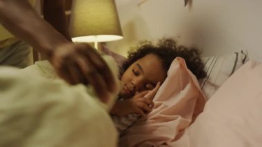 Geceleri rahat bir yatak odasında battaniyeli uyuyan Afro-Amerikan kıvırcık saçlı çocuğu izleyen tanınmamış bir babanın yan görüntüleri.
