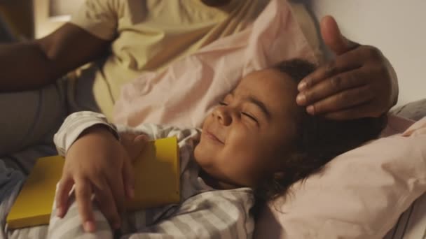 ベッドに横たわるアフリカ系アメリカ人の子供が黄色いカバーで本を抱擁しながら眠るために彼を置くために彼の頭の上にパット — ストック動画