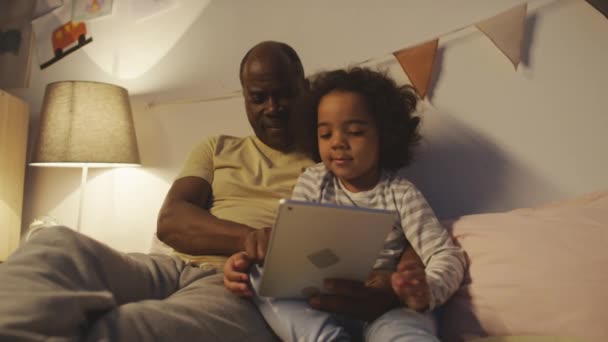 アフリカ系アメリカ人の父親がベッドに座ってベッドに座り 居心地の良いモダンなベッドルームでタブレットで見たり読んだりしながら抱きしめました — ストック動画