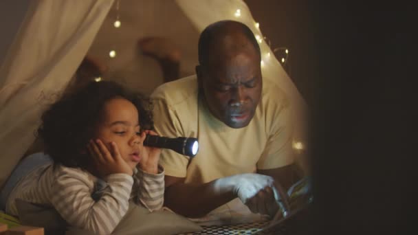 パジャマでアフリカ系アメリカ人の子供のパンニングショットは 夜に寝室で漫画を読んでいる父親とガーランドで飾られたフラッシュライトで横たわっています — ストック動画