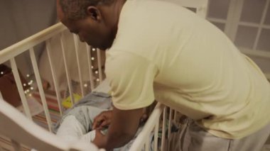 Çift ırklı küçük kızın bebek karyolasında ağlaması ve Afrikalı Amerikalı babanın onu kollarına alıp uyutup gece yatak odasında yatırması.