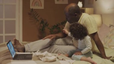 Afrika kökenli Amerikalı bir babanın evde iki ayaklı bir bebekle yatakta oturup bilgisayardan sağlık, kolik ve diş çıkarma hakkında video yoluyla kadın beyaz çocuk doktoruyla konuşmasının yan görüntüleri.