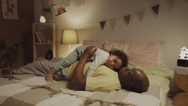 非洲裔美国人的父亲躺在床上 与生小孩的女婴躺在床上 晚上在床上休息 同时哄着蹒跚学步的幼儿在卧室里睡觉 — 图库视频影像