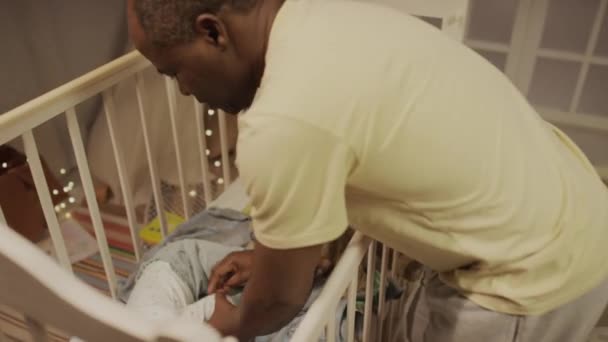 ベビーコットとアフリカ系アメリカ人の父親で泣いているBiracial幼児の側近は 夜寝室で寝るために赤ん坊を服を着た — ストック動画