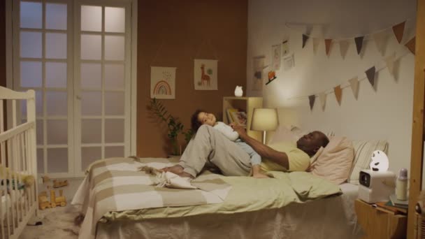 ベッドに横たわるアフリカ系アメリカ人の父親のサイドフルショットと 夜に装飾された寝室で彼の周りで寝るために2歳の赤ん坊の少女を暗殺 — ストック動画