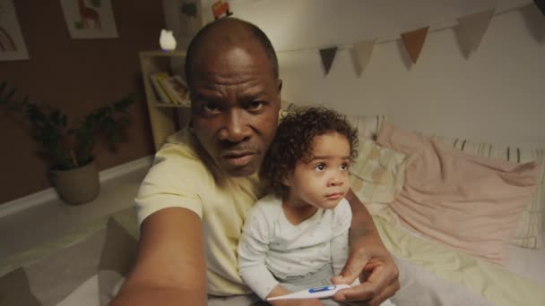 教资会拍摄的非洲裔美国人父亲拿着体温计 一边坐在床上 一边看着屋里的摄像机 一边谈论处方药 — 图库视频影像