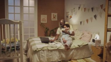 Akşamları dekore edilmiş yatak odasında iki yaşındaki iki yaşındaki melez kızı beslerken Afro-Amerikan ebeveynin yatağında dizüstü bilgisayarda çalışırken çekilmiş tam bir görüntüsü.