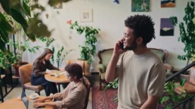 Orta boy polyetik veya gözlüklü Afro-Amerikan bir adamın şık bir kafeteryada dikilip cep telefonuyla konuşurken ve arka planda çalışan kadın müşterilerin fotoğrafları.