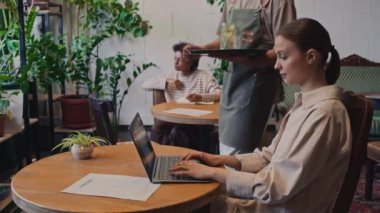 Kafe veya restoranda oturan, dizüstü bilgisayarda yazan, garsonun bir bardak limonatasını getirdiği, gülümseyen, teşekkür eden ve içki içen orta boy beyaz bir kadın resmi.