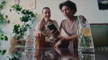 Kafkasyalı ve Afrikalı Amerikalı kız arkadaşların kafeteryada birlikte oturup, akıllı telefondaki fotoğraflara bakıp, erkek arkadaşlarını tartışırken, kafa sallarken ve limonata içerken orta seçici bir odak noktası.