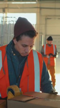 Çift ırklı depo erkek işçisinin iş günü geniş bir depoda koli bandıyla kutuyu kapatırken dikey görüntüsü.