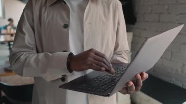 Orta yakın çekim, sakallı, bej gömlekli gülümseyen Afro-Amerikan bir adamın ofis tesisinde dikilirken ve dizüstü bilgisayarında yazarken.