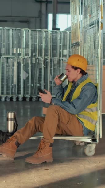 Biracial男性倉庫労働者がお茶やコーヒーを飲んで 在庫カートに座ってスマートフォンを使用し 職場での休憩中に笑顔で撮影したフルサイドショット — ストック動画