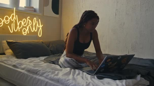 アフリカ系アメリカ人の若い女性がベッドに座り 現代のグラウンジロフトアパートで自宅からラップトップに取り組んでいる様子 — ストック動画