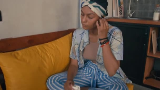 倾斜的中镜头 年轻的非洲裔美国妇女穿着耳塞坐在黄色的大胆沙发上 在家里用笔记本电脑工作 — 图库视频影像