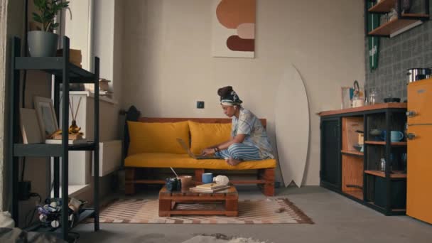 スタイリッシュなアフリカ系アメリカ人女性フリーランサーの全長映像がソファに座り モダンなロフトアパートでラップトップに取り組む — ストック動画