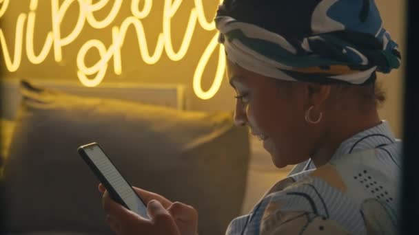 在带有霓虹灯题词的低调阁楼房子里 穿着班达纳手机聊天的快乐微笑的年轻非洲裔美国女人的侧面特写 — 图库视频影像