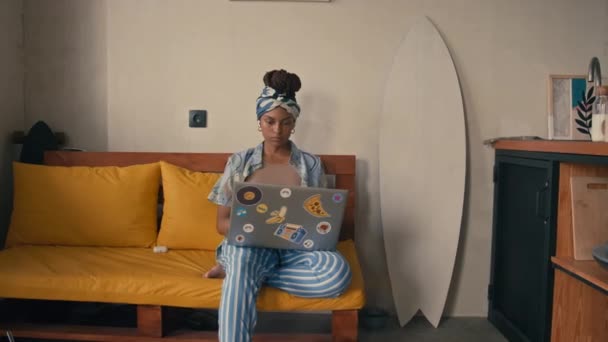 家から遠隔操作で働く若いアフリカ系アメリカ人女性の完全な肖像画 現代の家で黄色いソファーに座っているカメラのためのノートパソコンと笑顔 — ストック動画