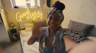 Bandanalı ve şık giyinen gülümseyen Afro-Amerikan bayanın orta sınıf POV UGC 'si mutlu bir şekilde kameraya bakıyor ve grunge evinde blog için video kaydediyor. Üzerinde sadece Good Vibes yazıyor.