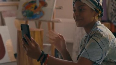 Bandana 'da gülümseyen Afrikalı Amerikalı genç modaya uygun bir bayanın video görüntüsü ya da video kaydı ile modern odada resimli akıllı telefona bakarken.