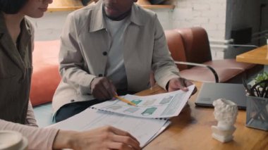 Afrika kökenli Amerikalı genç bir adamın ortak ofis tesisinde koltukta oturup, mali raporlardaki grafiklere, verileri açıklarken ve kadın meslektaşının dinlediği ve kafa salladığı orta ölçekli yakın çekim.