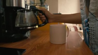 Tanımlanamayan bir kadının kahve makinesinden fincana kahve dökerek eve gidişinin yan görüntüsü.