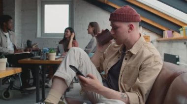 Bereli genç bir beyaz adamın iş yerinde koltukta otururken, akıllı telefon kullanıp kahve içerken ve çeşitli girişim takımlarının arka planda iş görüşmesi yaparken orta boy fotoğrafı.