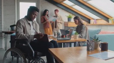 Özürlü Afrikalı Amerikalı bir adamın orta boy portresi. Tekerlekli sandalyede, ortak ofis tesisinde oturuyor. Defteri kapatıyor ve arka planda çalışan diğer takım üyelerine bakıyor.