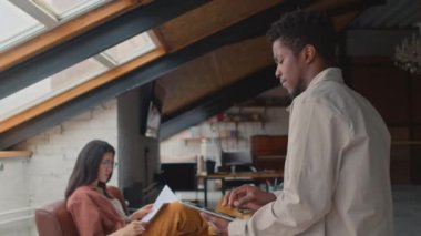 Orta boy Afro-Amerikan bir adamın çatı katında çalışma odasında tablet bilgisayar kullanırken, sonra çatı penceresine dalgın dalgın bakarken ve arka planda broşürle çalışan Asyalı bir kadının fotoğrafı.