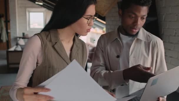 持有证件的年轻亚洲妇女和携带手提电脑的非洲裔美国男子在合作办公室里站在一起 观看屏幕并讨论一些工作问题的中等特写镜头 — 图库视频影像