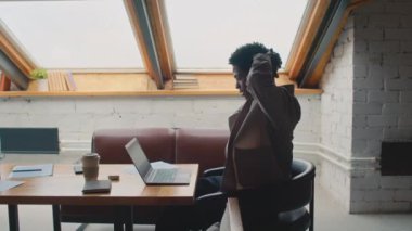 Orta boy Afro-Amerikan bir adamın sandalyede otururken, laptopta çalışırken, ofisini paylaşırken, sonra ellerini kafanın arkasına yapıştırırken, gerinirken, yanlara doğru eğilirken ve yazı yazmaya devam ederken.