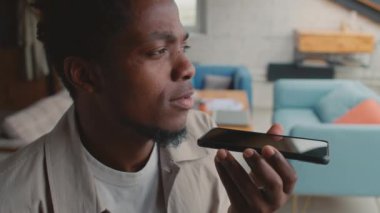 Afrika kökenli Amerikalı genç bir adamın iş yerinde otururken, akıllı telefonu yatay olarak tutarken, düşünceli bakışlar atarken ve mikrofona sesli mesaj kaydederken yakın plan görüntüsü.