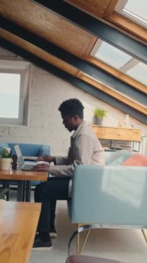 Full vertical shot of busy young African American male freelancer in casual clothes sitting alone at table on couch in loft coworking office, looking at document and typing on laptop
