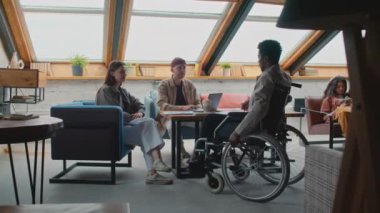 Full shot of African American man in wheelchair joining colleagues at team meeting in coworking office, greeting and submitting document draft to manager, Asian female customer sitting with smartphone