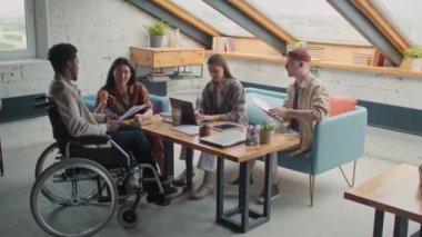 Full shot of multiracial project team working together in loft office - Asian woman explaining idea to black man in wheelchair, and Caucasian female and male colleagues with laptop discussing proposal