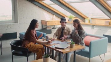 Full shot of Caucasian man and two Asian female colleagues sitting at table and discussing business proposal draft with financial charts, black guy with laptop walking in and joining team meeting