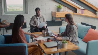 Medium zoom-in of African American man with disability sitting in wheelchair at startup team meeting, proposing new presentation design, female colleagues watching and listening