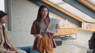 Medium zoom out shot of multiethnic startup team having meeting in loft coworking - Asian female manager displaying financial graph and reporting results, colleagues asking questions and arguing